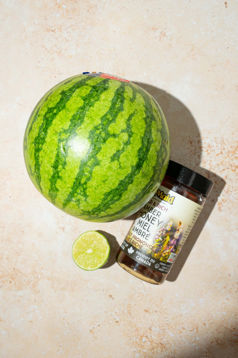 A tan counter with ingredients to make watermelon shaved ice including a whole watermelon, honey, and lime.