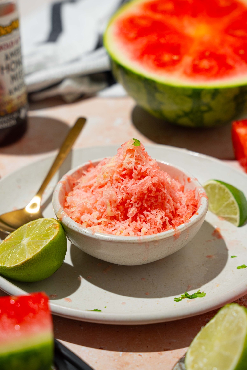 A small white bowl with watermelon shaved ice drizzled with honey. The bowl is on a white plate with a spoon and lime. The plate is on a tan counter with a watermelon.