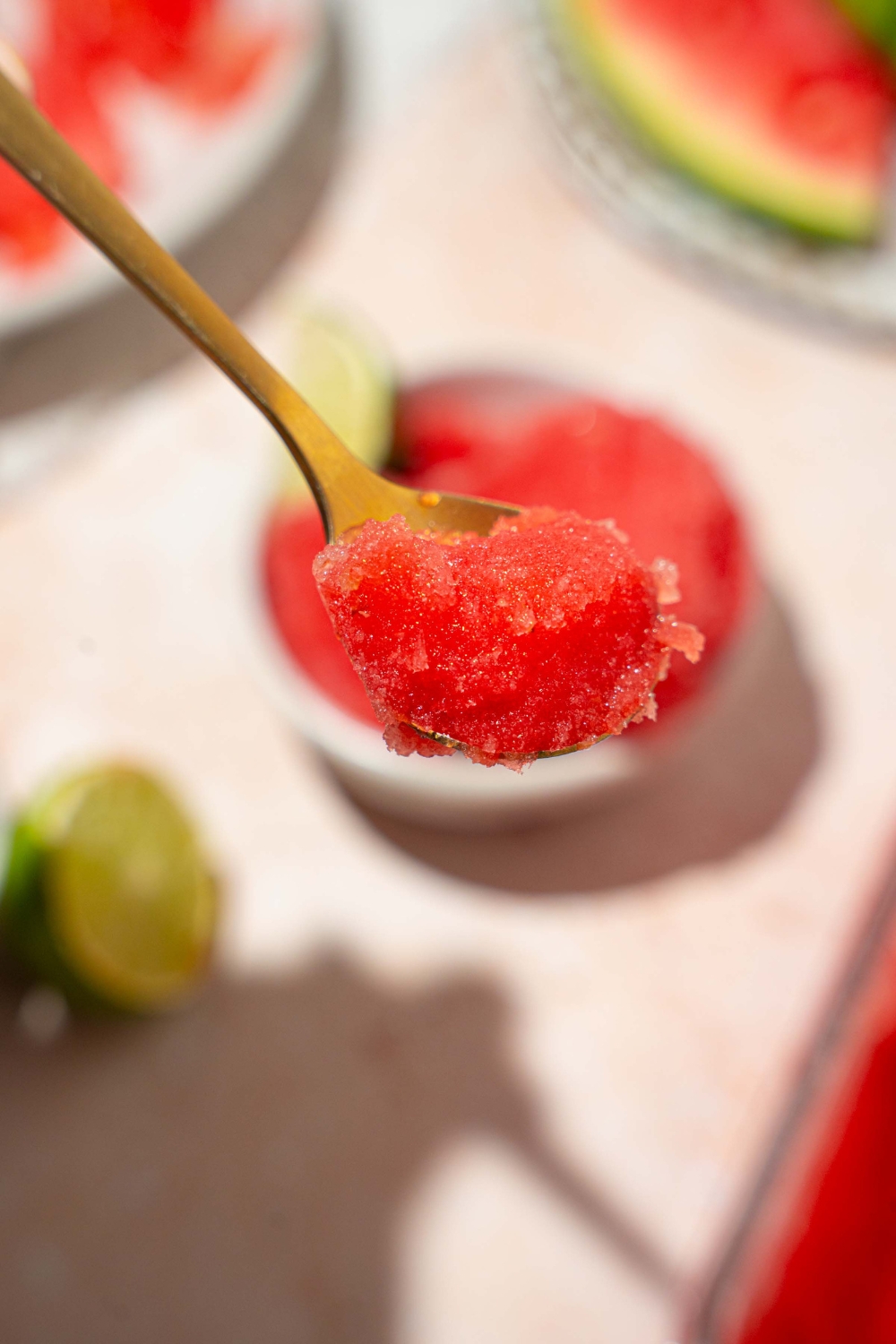 A close up of a spoon with a bite of watermelon sorbet. There is a bowl of sorbet on a tan counter blurred in the background.