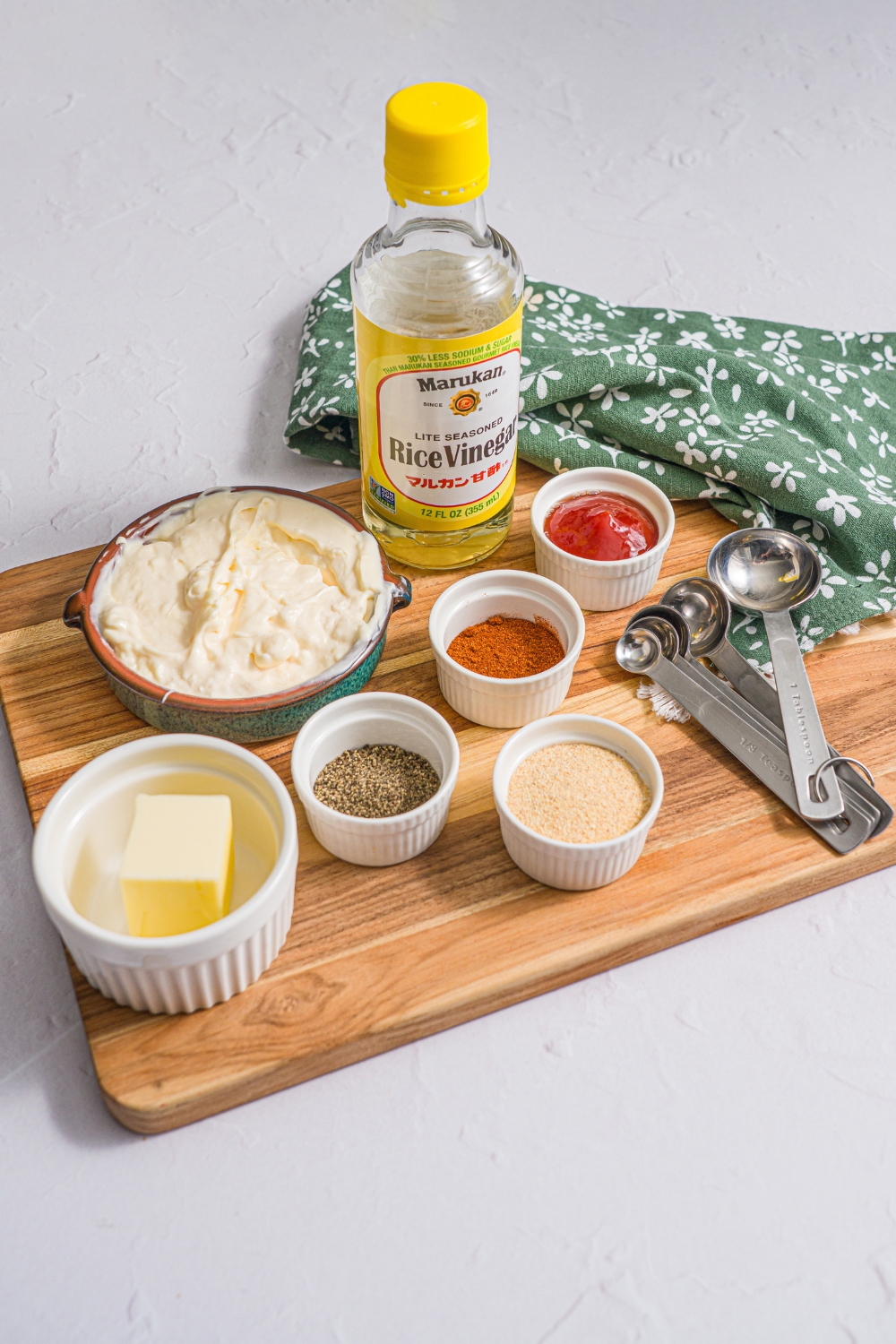 A wooden board with small bowls of ingredients containing ingredients to make yum yum hibachi sauce including mayo, ketchup, rice vinegar, butter, garlic powder, paprika, sugar, and black pepper. The board is on a white counter with a green floral napkin.
