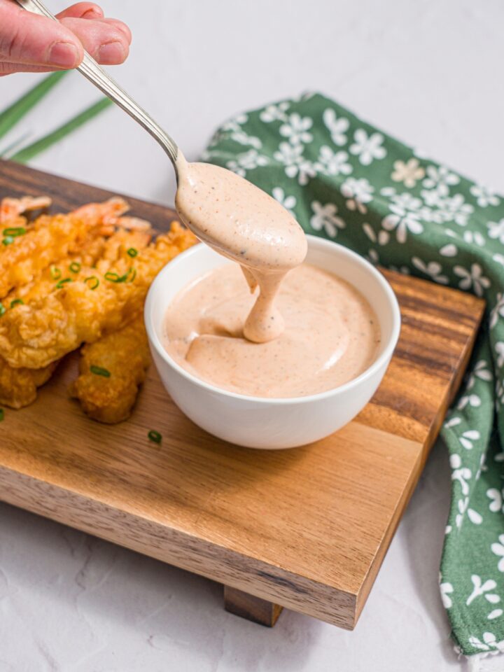 A wooden board with a bowl of yum yum hibachi sauce and crispy shrimp garnished with sliced green onion. A spoon is dipping into the bowl. The board is on a white counter with a green floral napkin.