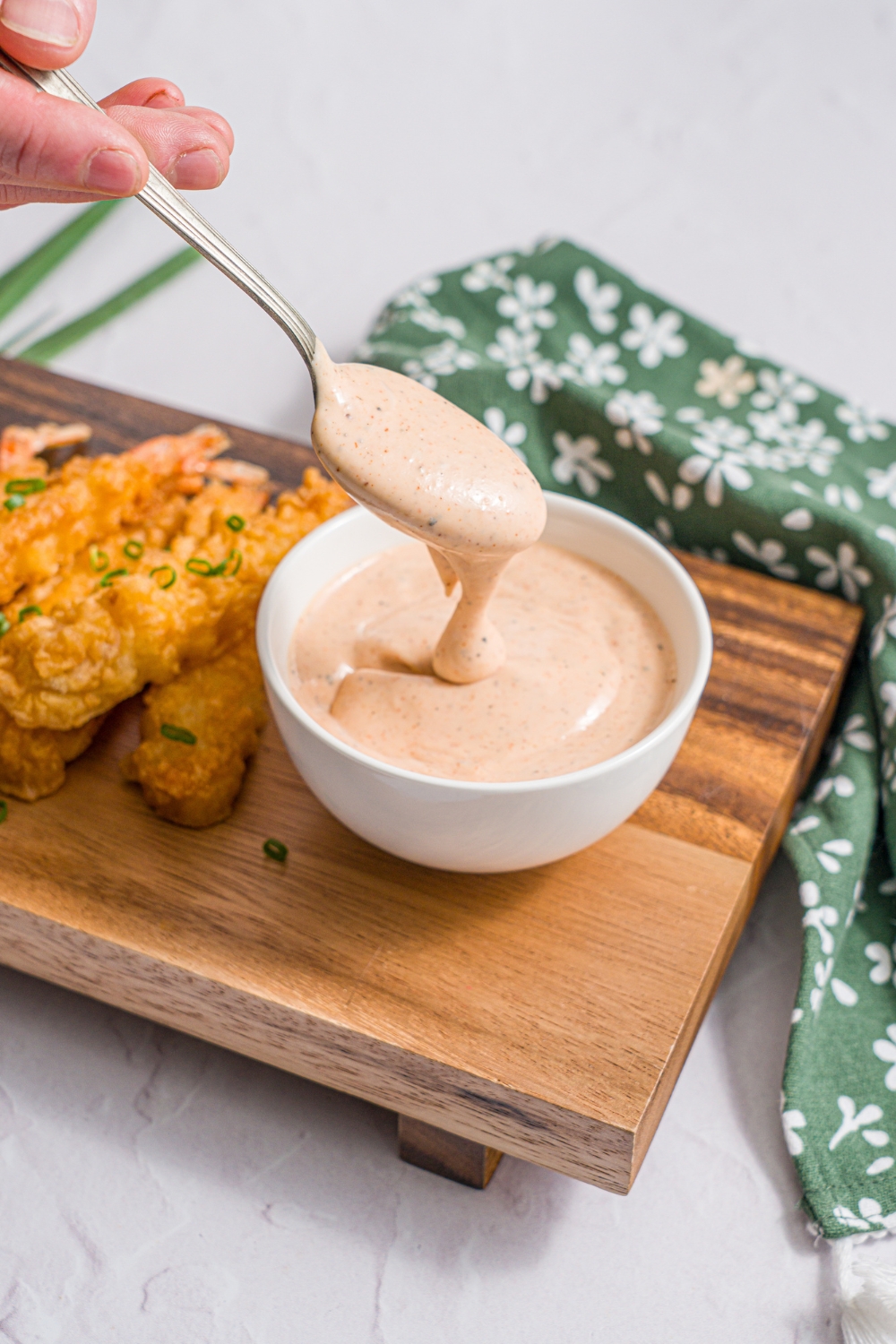 A wooden board with a bowl of yum yum hibachi sauce and crispy shrimp garnished with sliced green onion. A spoon is dipping into the bowl. The board is on a white counter with a green floral napkin.