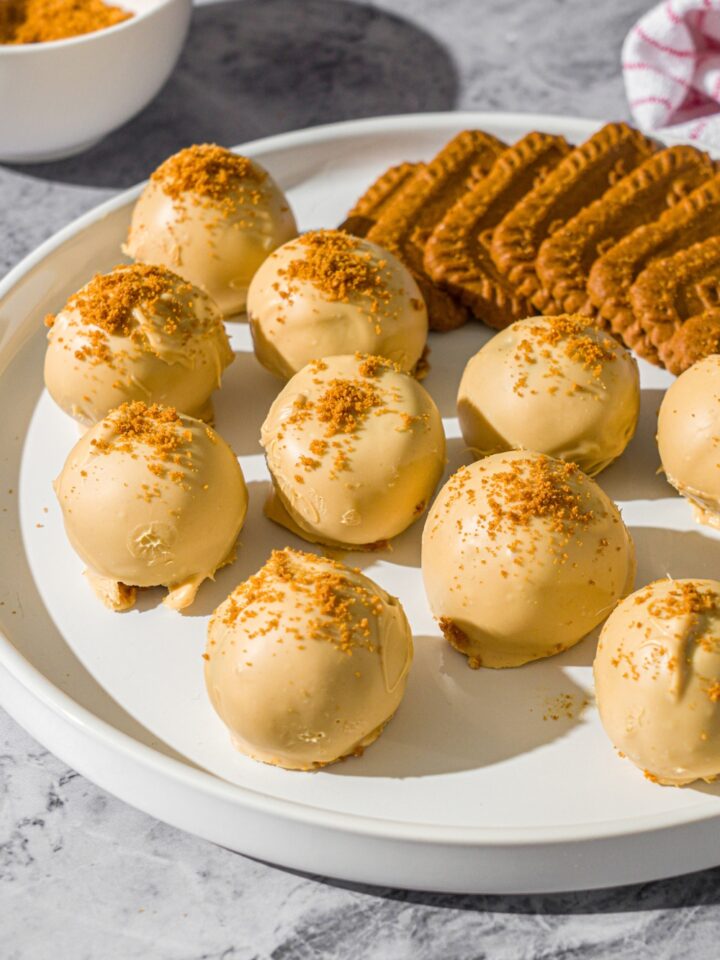 A white plate with Biscoff truffles coated in white chocolate and topped with cookie crumbs. . The truffles are served with Biscoff cookies. The plate is on a marble counter with a small bowl of cookie crumbs and red striped napkin.