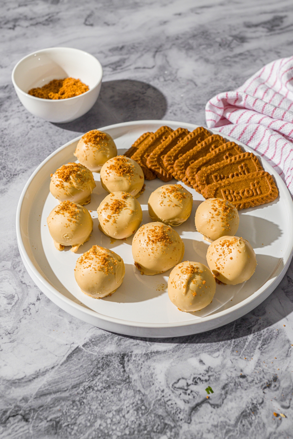 A white plate with Biscoff truffles coated in white chocolate and topped with cookie crumbs. The truffles are served with Biscoff cookies. The plate is on a marble counter with a small bowl of cookie crumbs and red striped napkin.