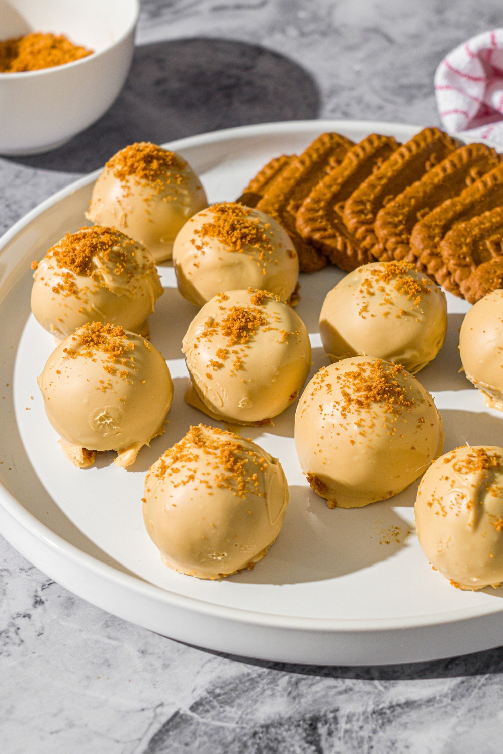 A white plate with Biscoff truffles coated in white chocolate and topped with cookie crumbs. . The truffles are served with Biscoff cookies. The plate is on a marble counter with a small bowl of cookie crumbs and red striped napkin.