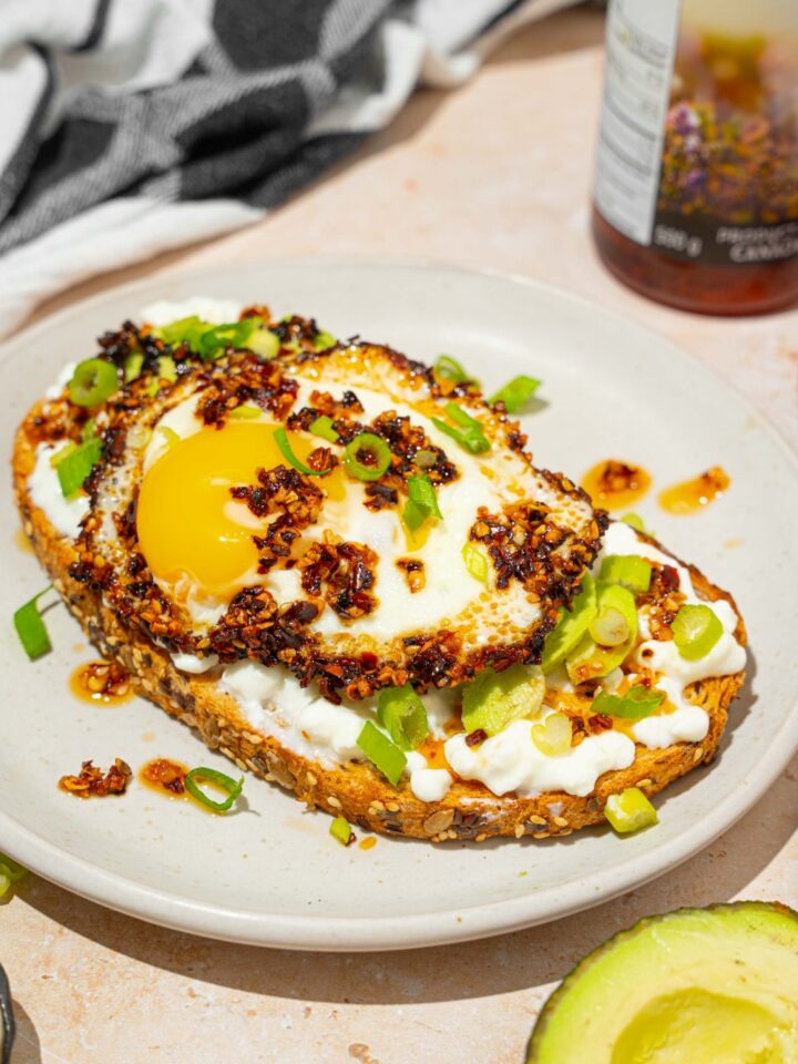 A white plate with a slice of chili crisp avocado toast garnished with sliced green onion. The plate is on a tan counter with a jar of chili crisp oil.