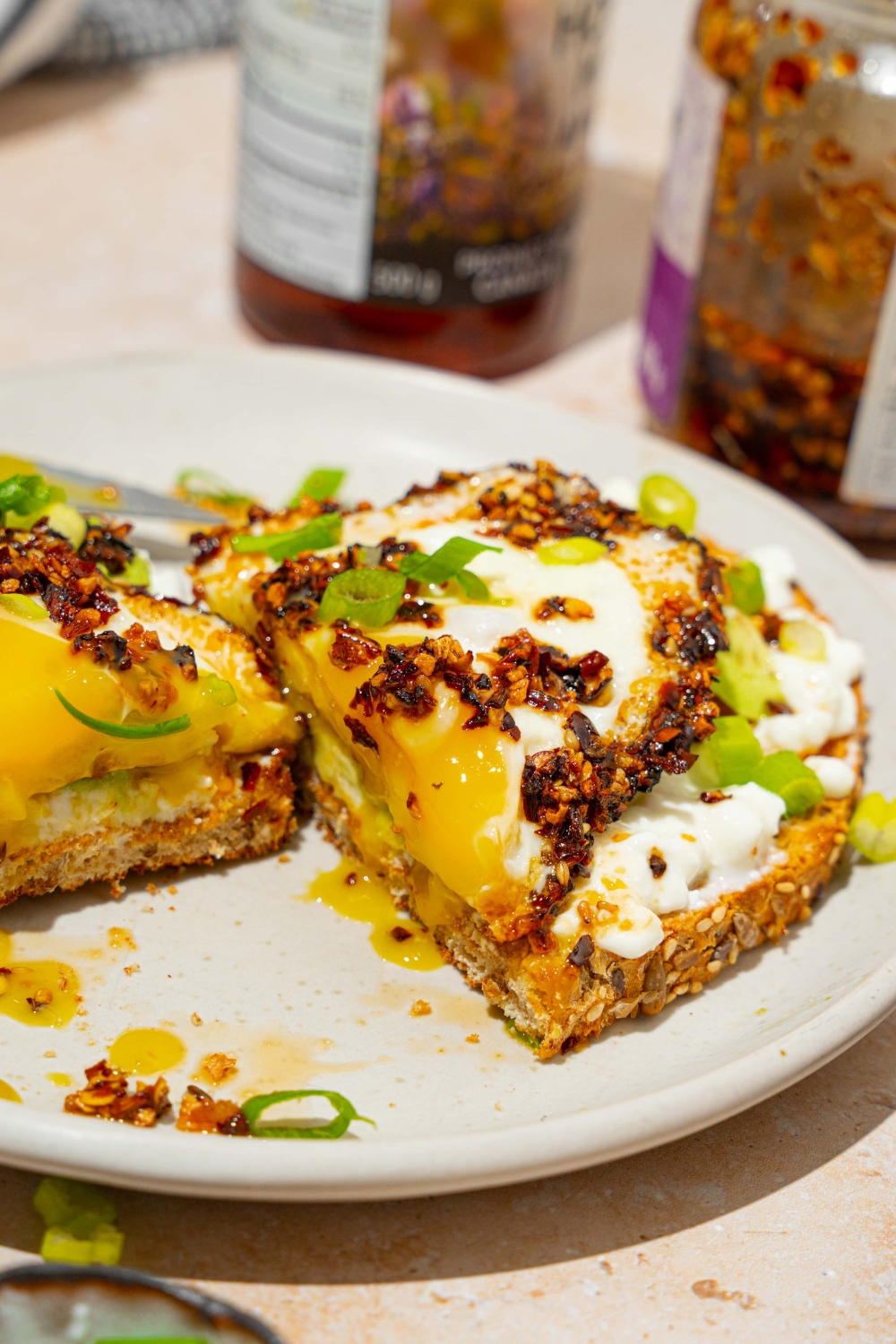 A white plate with a slice of chili crisp avocado toast garnished with sliced green onion. The slice of toast is sliced in half. The plate is on a tan counter with a jar of chili crisp oil.