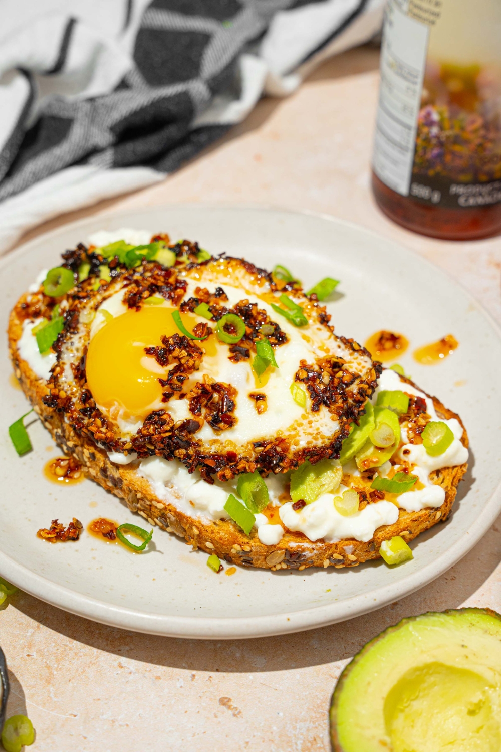A white plate with a slice of chili crisp avocado toast garnished with sliced green onion. The plate is on a tan counter with a jar of chili crisp oil.