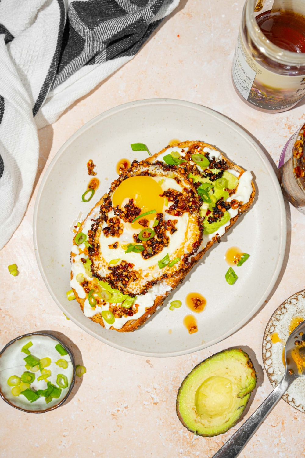 A white plate with a slice of chili crisp avocado toast garnished with sliced green onion. The plate is on a tan counter with a jar of chili crisp oil and avocado.