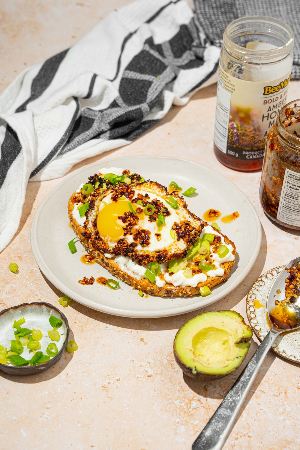A white plate with a slice of chili crisp avocado toast garnished with sliced green onion. The plate is on a tan counter with a jar of chili crisp oil and avocado.