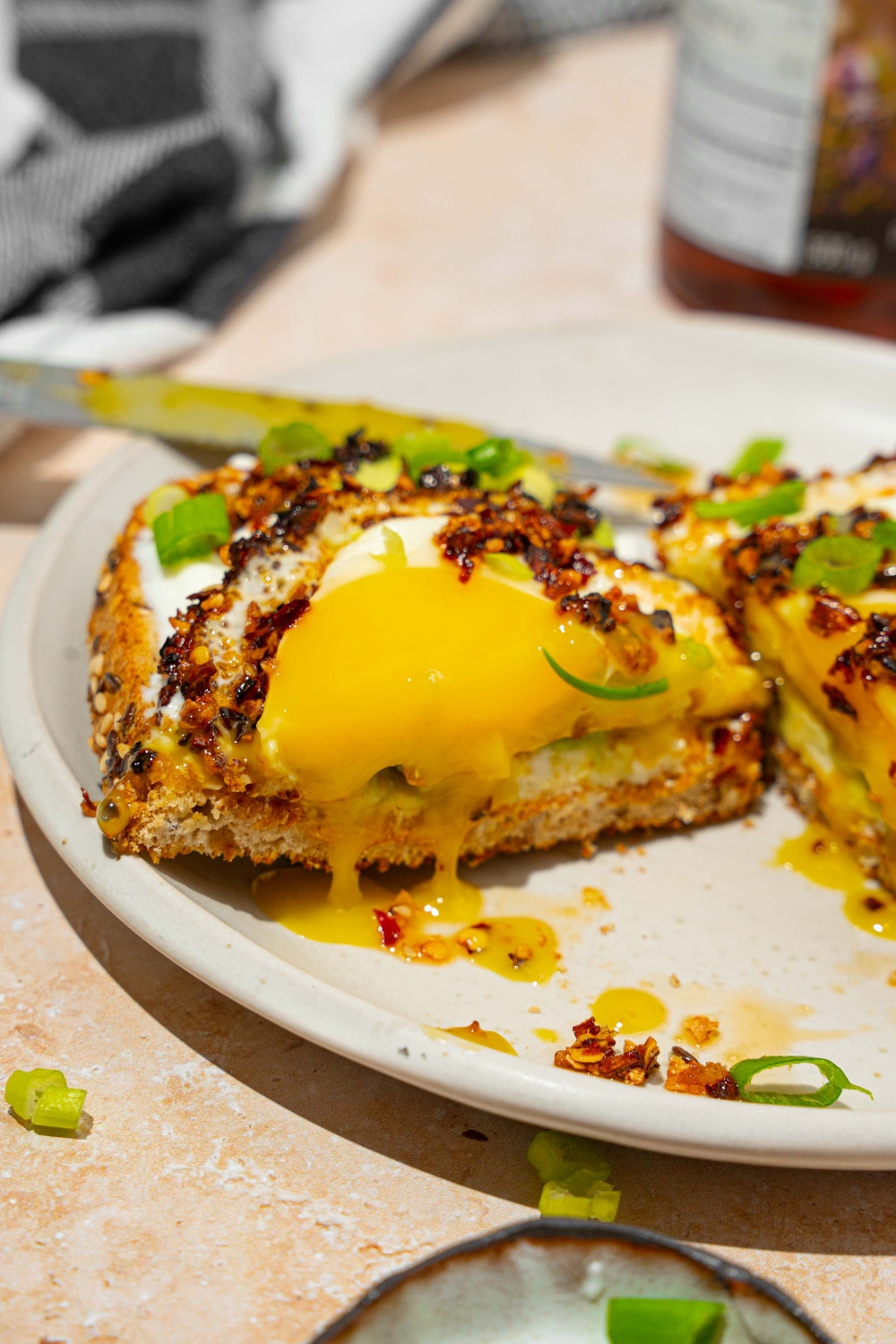A white plate with a slice of chili crisp avocado toast garnished with sliced green onion. The toast is sliced in half. The plate is on a tan counter with a jar of chili crisp oil.