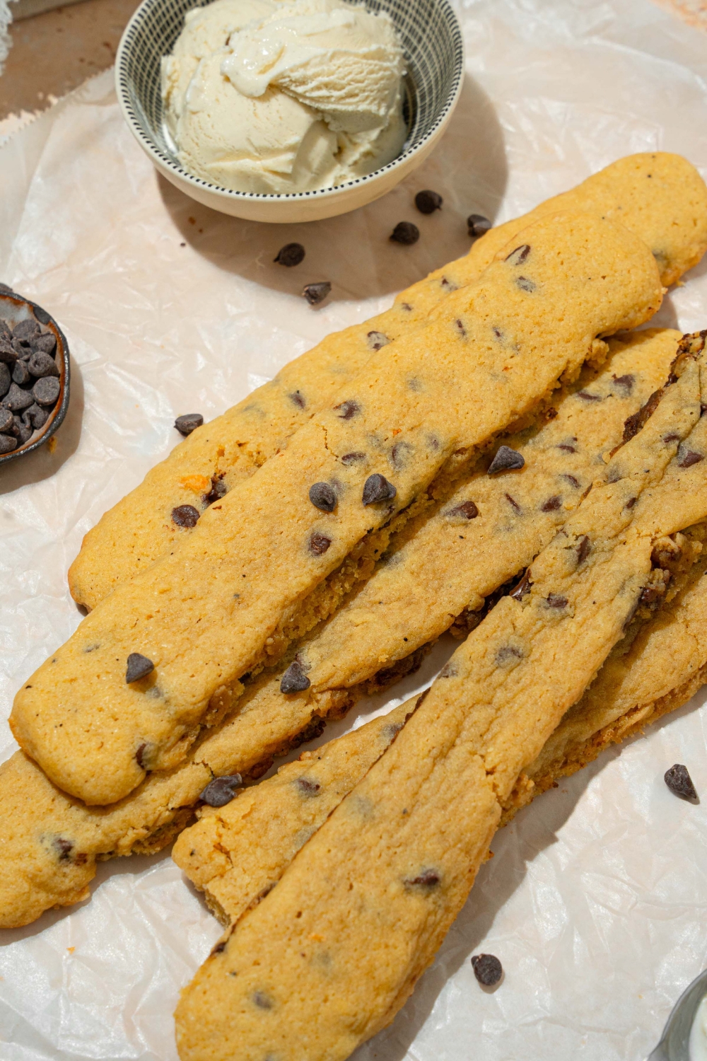 Several chocolate chip cookie fries on a piece of parchment paper sprinkled with chocolate chips. There is a bowl of vanilla ice cream.
