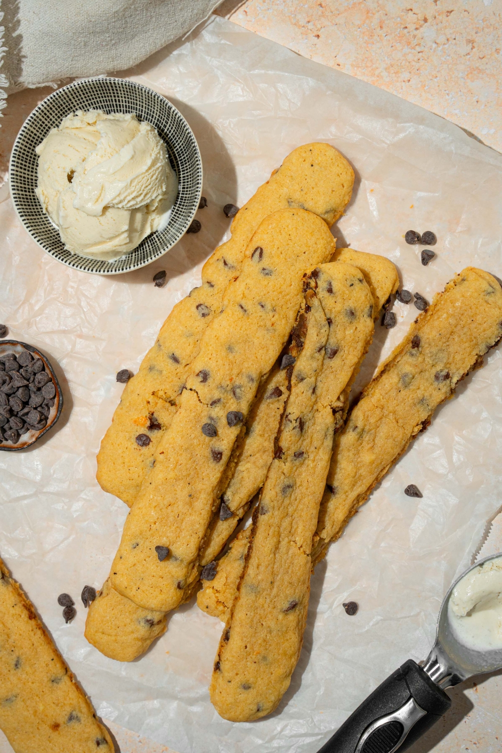 Several chocolate chip cookie fries on a piece of parchment paper sprinkled with chocolate chips. There is a bowl of vanilla ice cream.