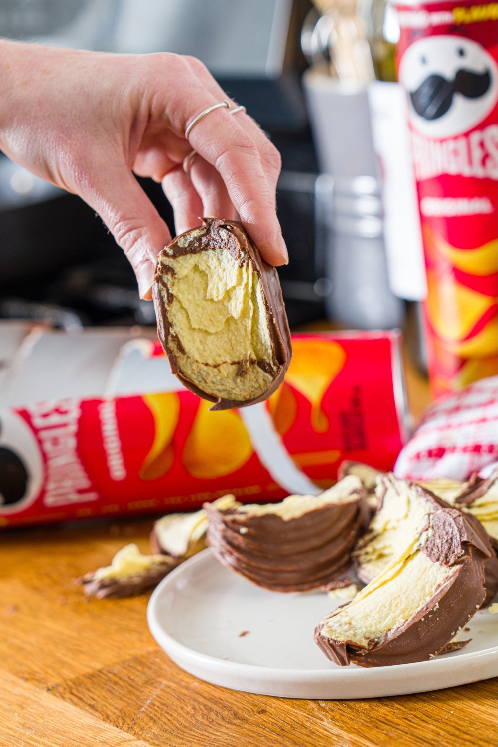 A hand holding a piece of a Pringles chocolate block over a white plate with some more pieces of the Pringles chocolate on it.