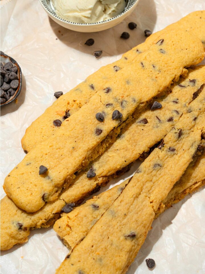 A bunch of chocolate chip cookie fries on a sheet of parchment paper with part of a bowl of vanilla ice cream behind it.