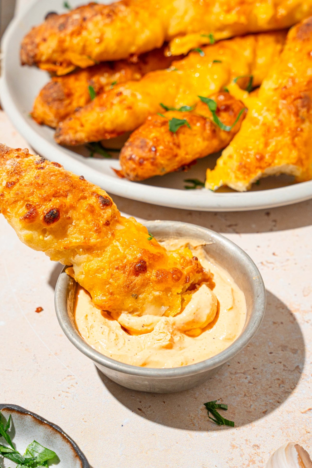 A cottage cheese breadstick dipped in a small cup of dipping sauce. The cup is on a tan counter with a plate of breadsticks.