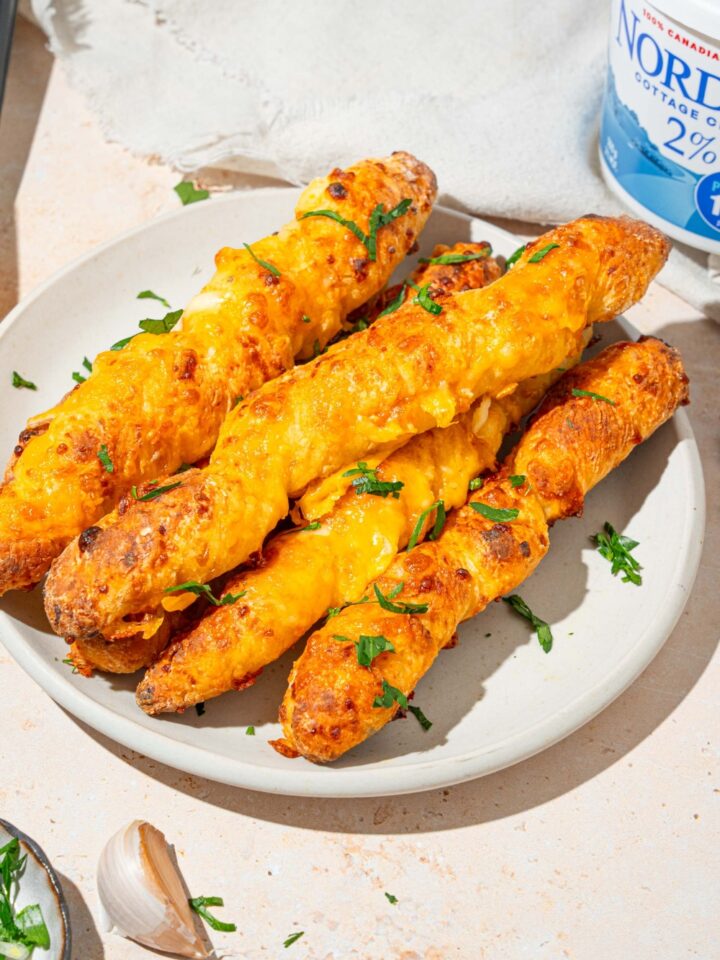 A white plate with a pile of cottage cheese breadsticks garnished with fresh parsley. The plate is on a tan counter with garlic and a white cloth napkin.
