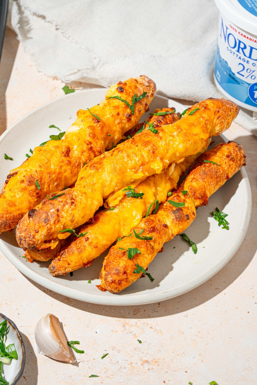 A white plate with a pile of cottage cheese breadsticks garnished with fresh parsley. The plate is on a tan counter with garlic and a white cloth napkin.