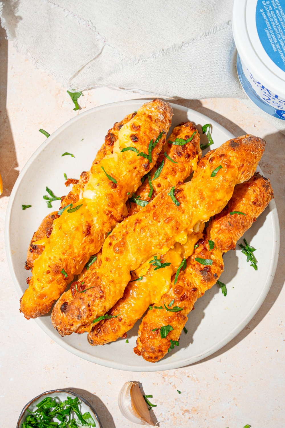 A white plate with a pile of cottage cheese breadsticks garnished with fresh parsley. The plate is on a tan counter with garlic and a white cloth napkin.