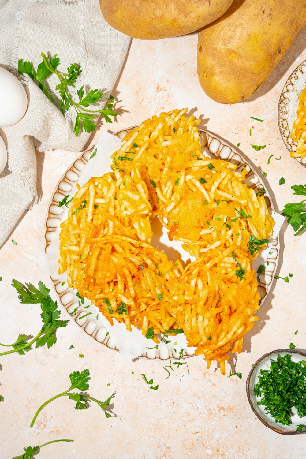 A plate lined with parchment paper with crispy parmesan hashbrowns garnished with fresh parsley. The plate is on a tan counter with plate of garnishes and a white cloth napkin.