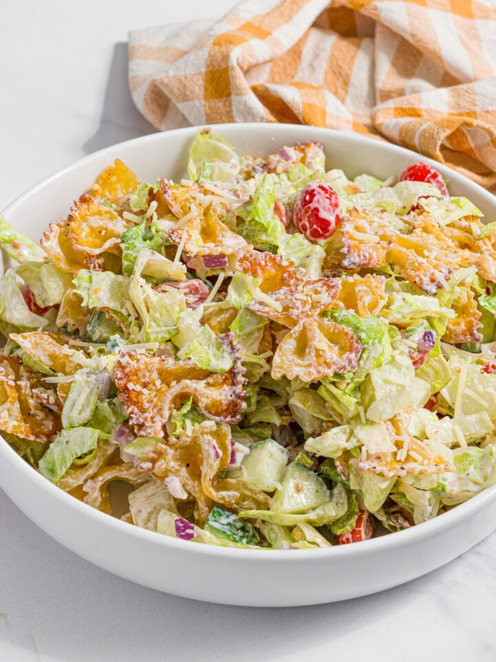 A white bowl with crispy pasta salad tossed with shredded lettuce, tomatoes, cucumber, and onion in a yogurt dressing. The bowl is on a marble counter with a yellow checkered napkin.
