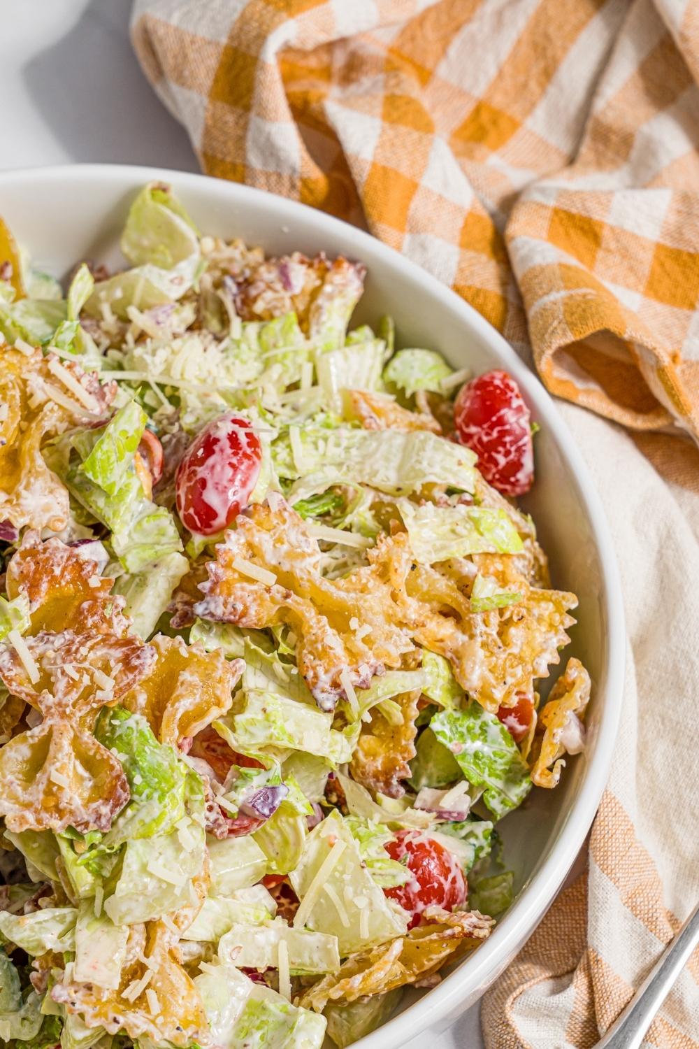 A white bowl with crispy pasta salad tossed with shredded lettuce, tomatoes, cucumber, and onion in a yogurt dressing. The bowl is on a marble counter with a yellow checkered napkin.