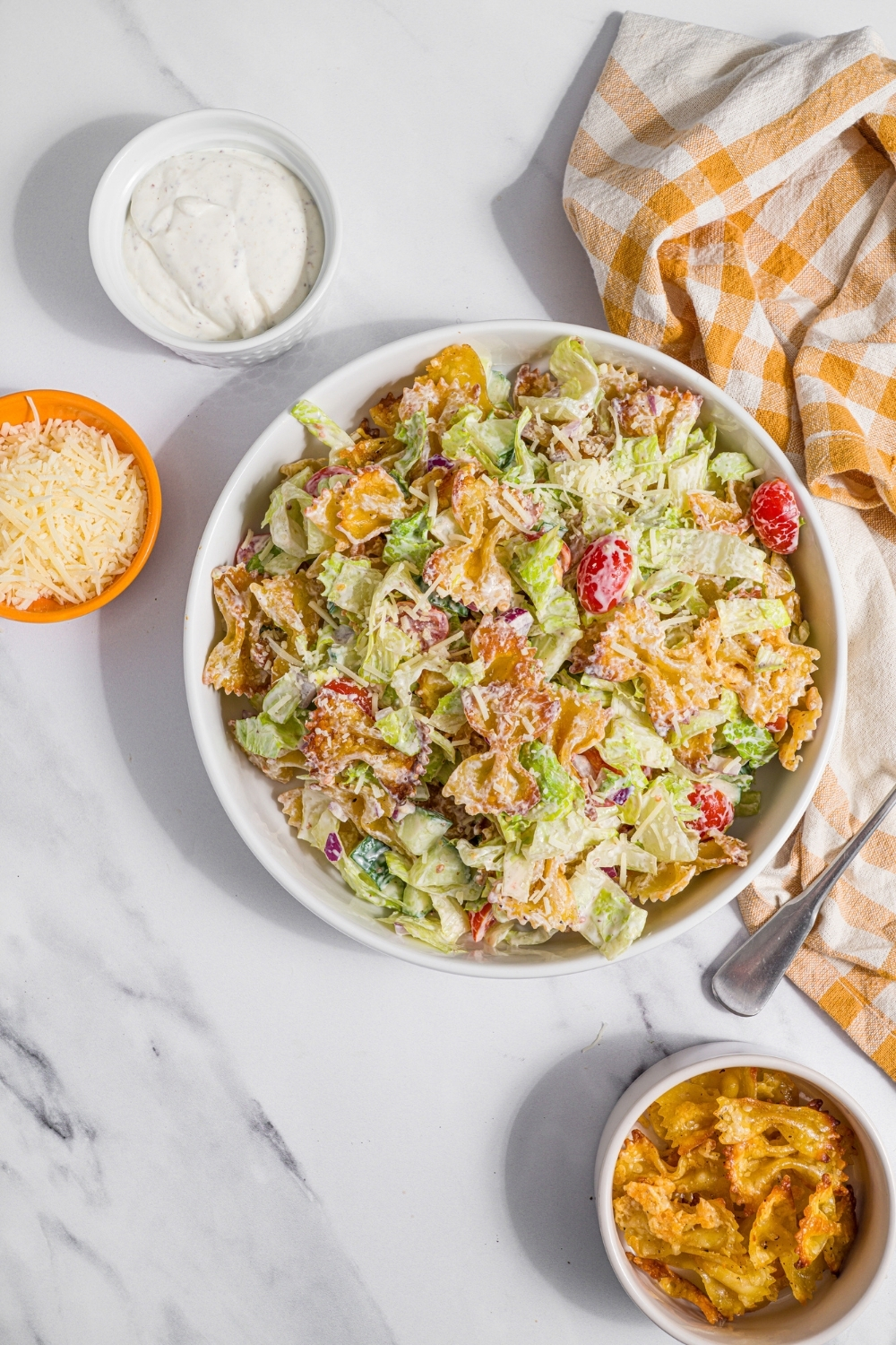 A white bowl with crispy pasta salad tossed with shredded lettuce, tomatoes, cucumber, and onion in a yogurt dressing. The bowl is on a marble counter with a yellow checkered napkin and small bowls of ingredients.