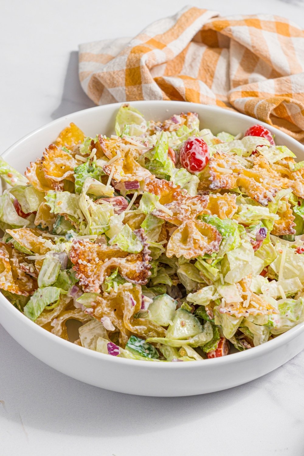 A white bowl with crispy pasta salad tossed with shredded lettuce, tomatoes, cucumber, and onion in a yogurt dressing. The bowl is on a marble counter with a yellow checkered napkin.