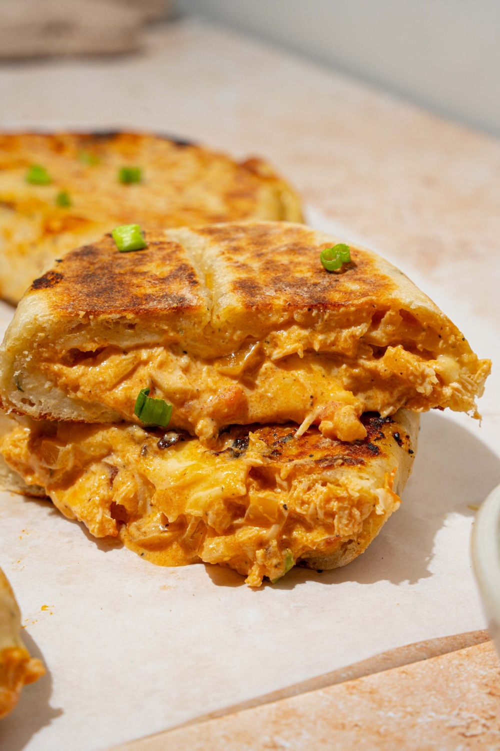 A piece of parchment paper with several Buffalo chicken hot pockets garnished with chives. One of the hot pockets is sliced in half and stacked on one another. The paper is on a tan counter with a bowl of dipping sauce.