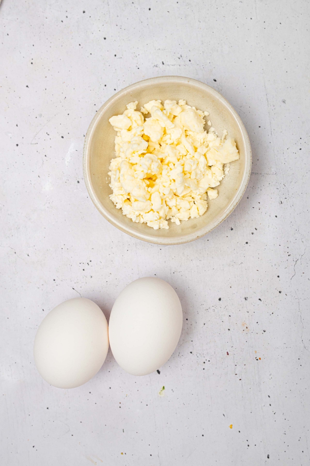 A bowl of feta cheese and two eggs on a white counter.