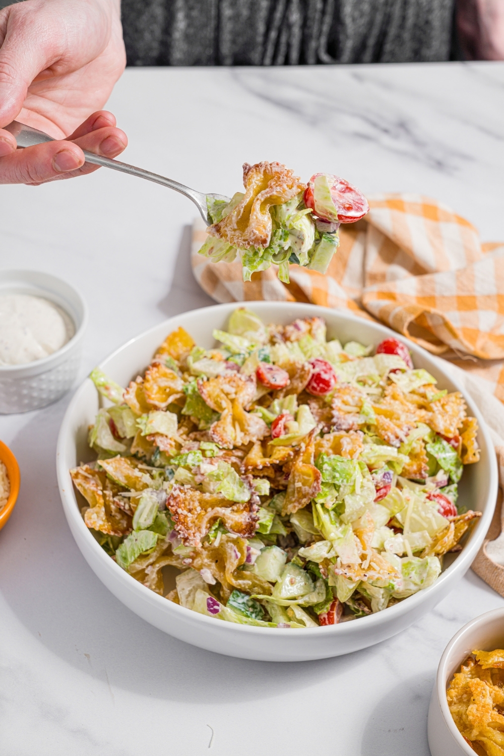A white bowl with crispy pasta salad tossed with shredded lettuce, tomatoes, cucumber, and onion in a yogurt dressing. There is a fork with a bite of pasta salad over the bowl. The bowl is on a marble counter with a yellow checkered napkin.