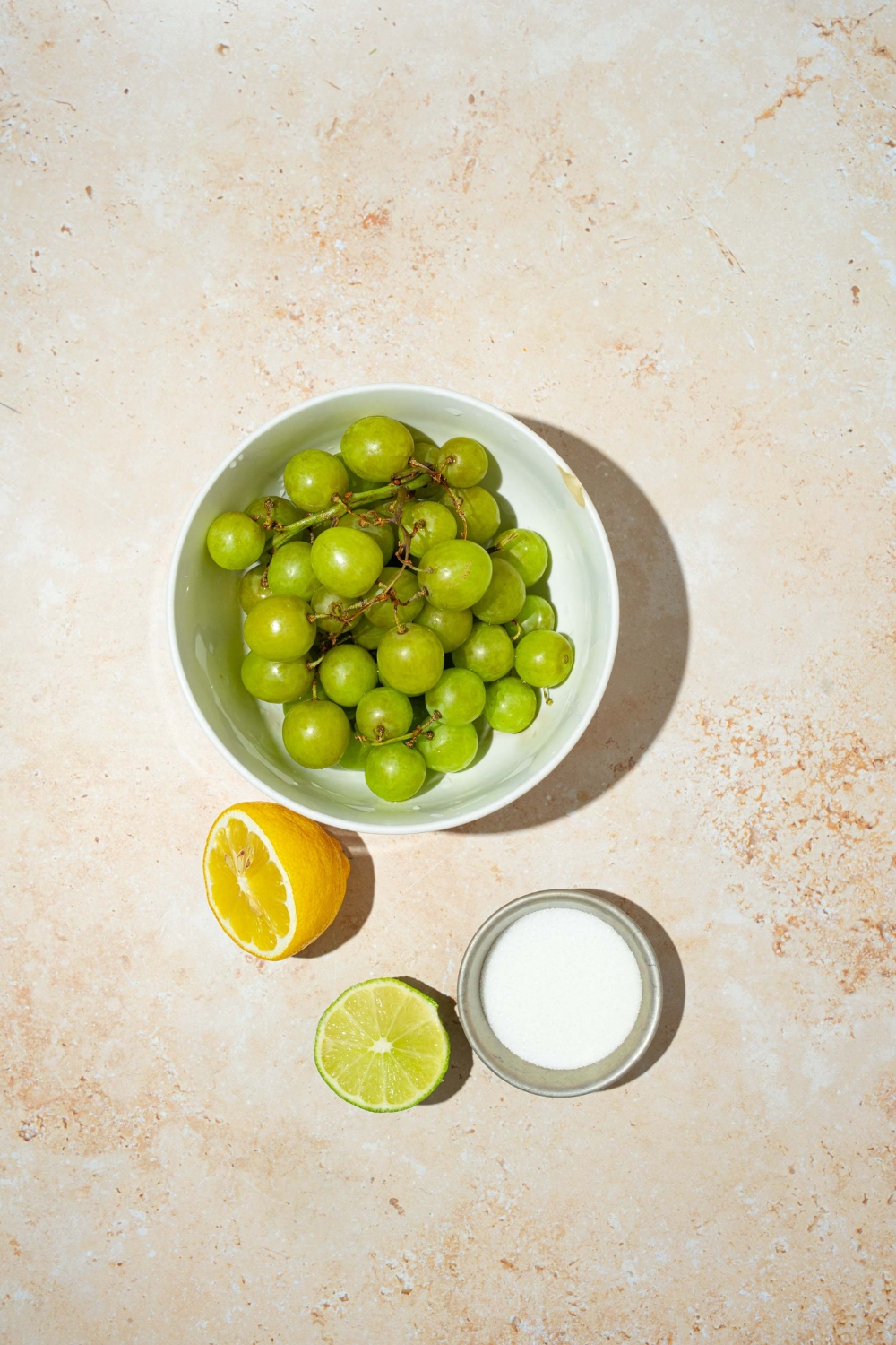 A bowl of green grapes, a bowl of sugar, a half of a lemon, a half of a lime on a white counter.