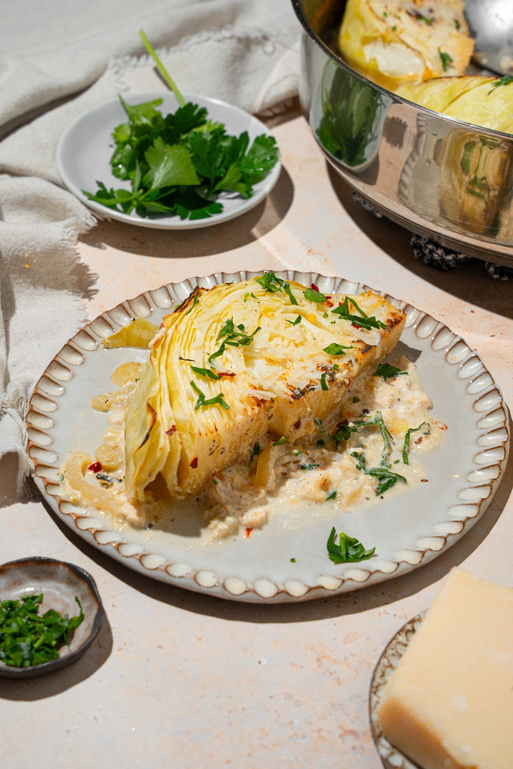 A plate with a slice of garlic parmesan melted cabbage garnished with fresh parsley. The plate is on a tan counter with a small bowl of garnishes and parmesan cheese wedge.