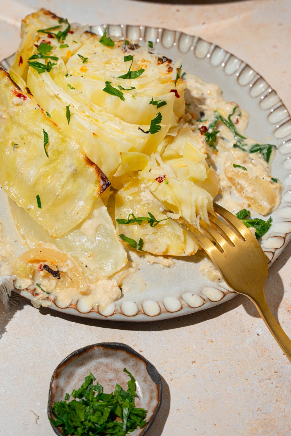 A plate with a slice of garlic parmesan melted cabbage garnished with fresh parsley. A fork is taking a bite of cabbage. The plate is on a tan counter with a small bowl of garnishes.