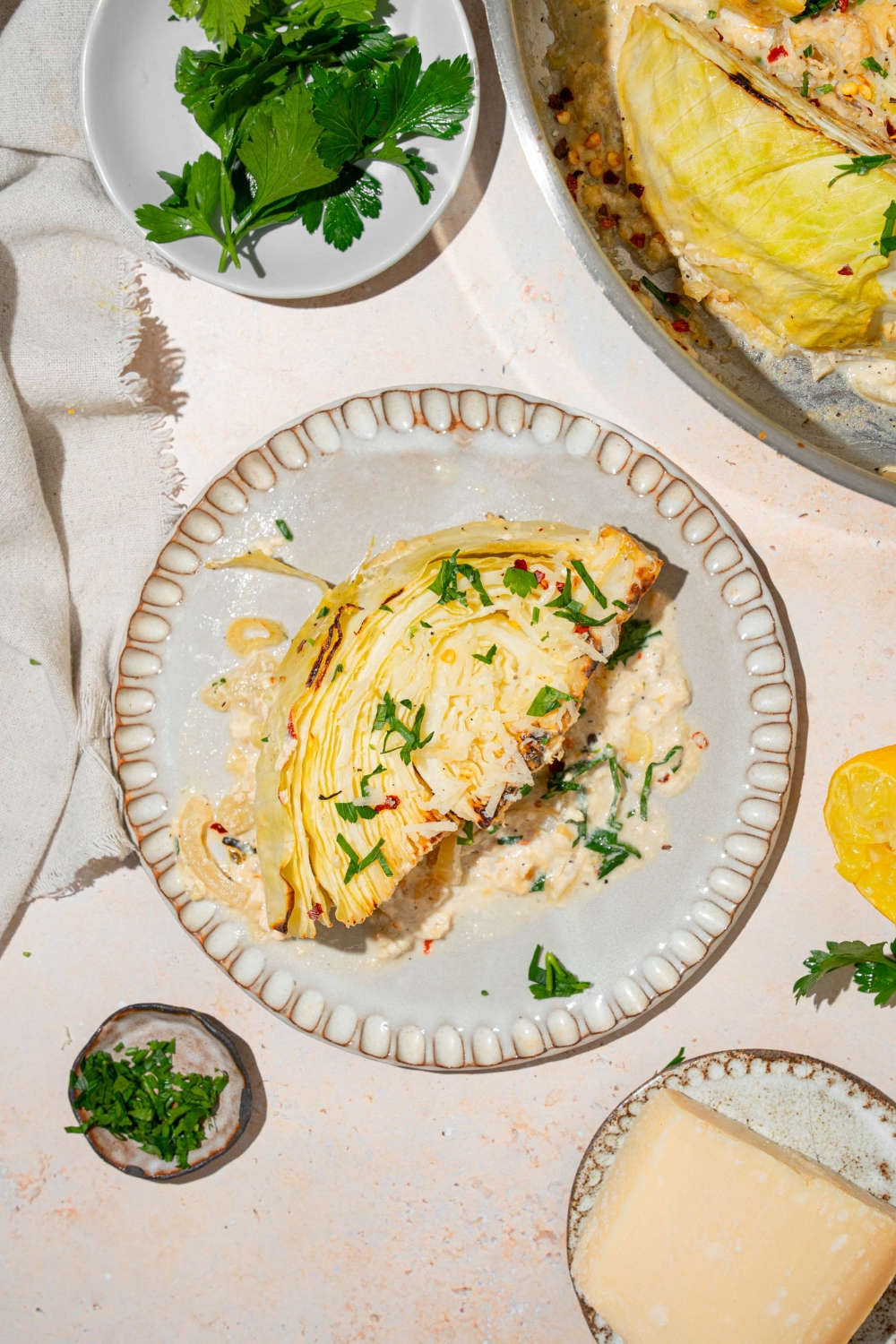 A plate with a slice of garlic parmesan melted cabbage garnished with fresh parsley. The plate is on a tan counter with a small bowl of garnishes, parmesan cheese wedge, and skillet of melted cabbage.