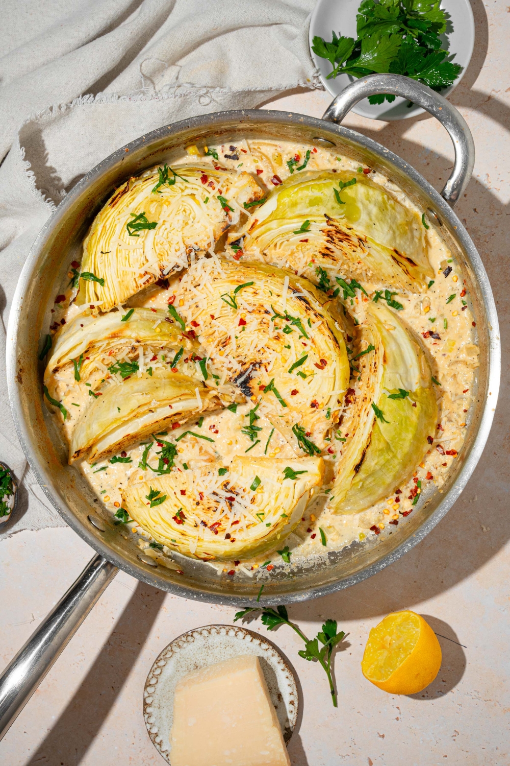 A skillet with baked garlic parmesan melting cabbage garnished with fresh parsley, red pepper flakes, and shredded parmesan cheese. The skillet is on a tan counter with a white cloth napkin and bowls of garnishes.
