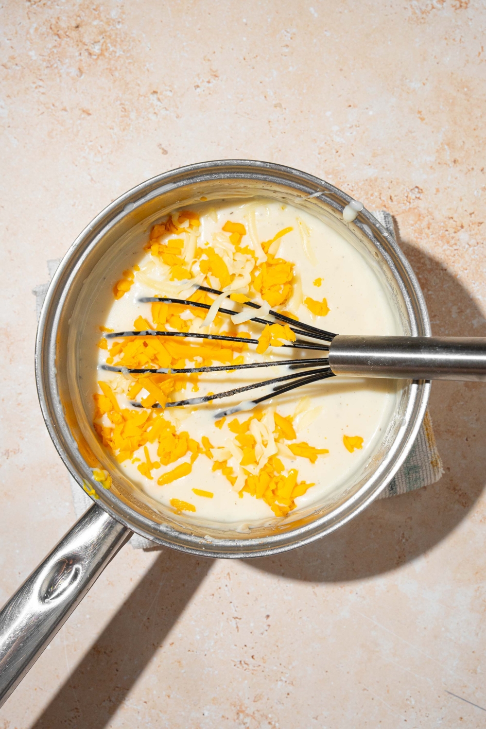 A whisk in a pot that is filled with a creamy sauce and shredded cheeses.