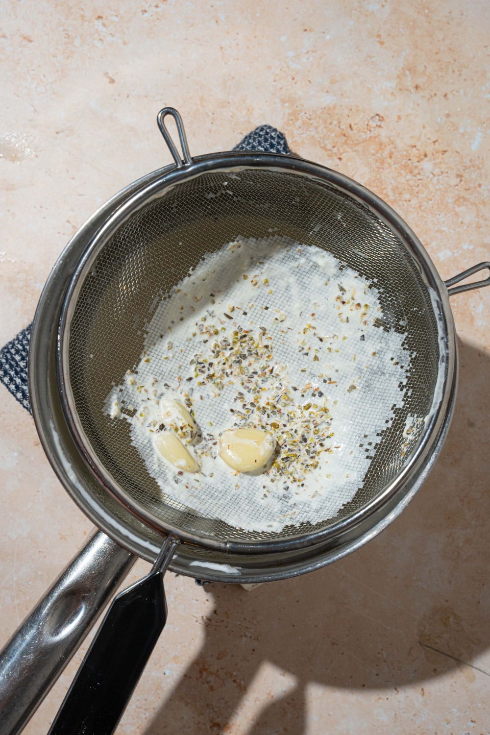 A pot with potato pave cream strained through a fine mesh strainer. Garlic and seasonings are remaining in the strainer. The pot is on a tan counter.