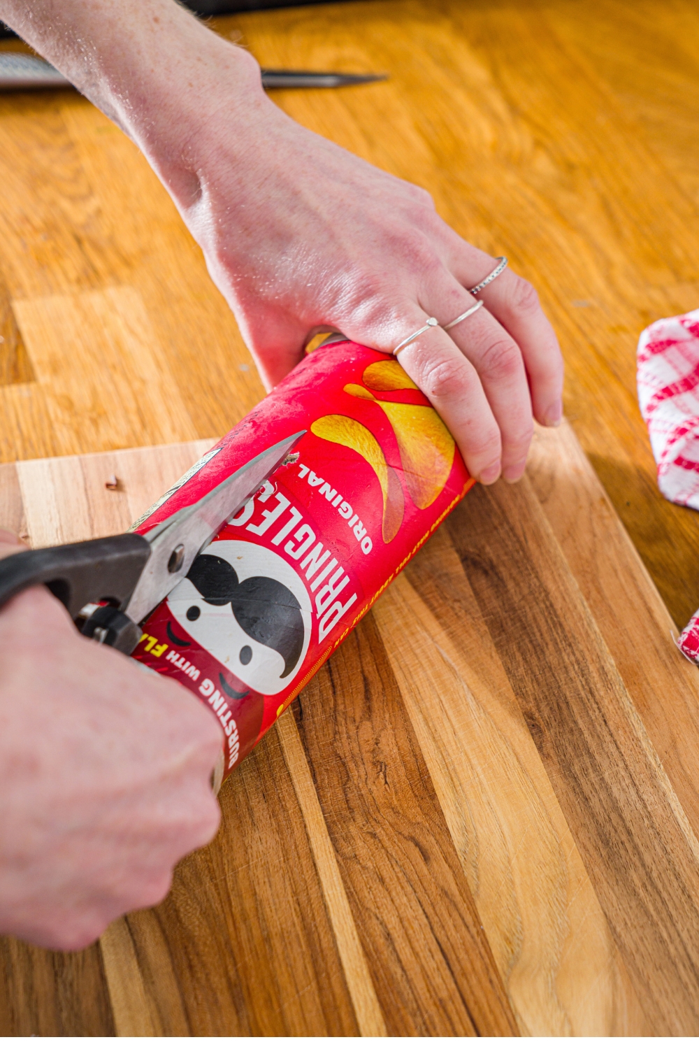 Two hands holding scissors and a Pringles can using the scissors to cut the can.
