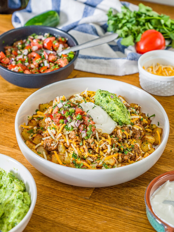 A white bowl with a loaded potato taco bowl including roasted potatoes, taco meat, shredded cheese, pico de gallo, sour cream, and guacamole. The bowl is garnished with cilantro. The bowl is on a wooden counter with bowls of toppings and a white striped napkin.