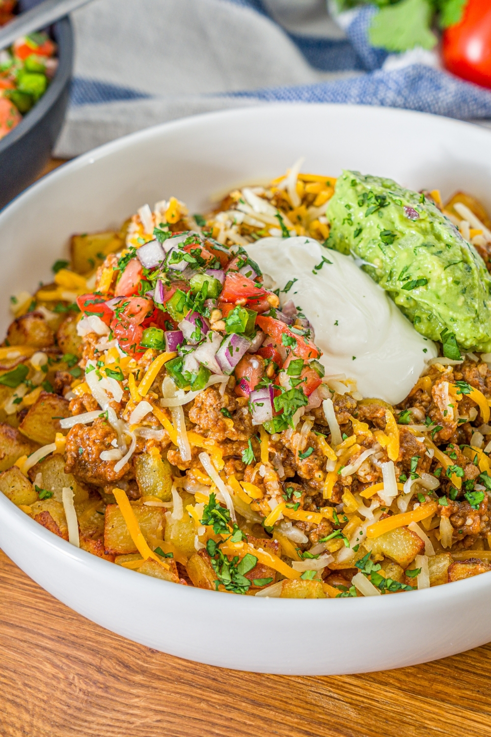 A white bowl with a loaded potato taco bowl including roasted potatoes, taco meat, shredded cheese, pico de gallo, sour cream, and guacamole. The bowl is garnished with cilantro. The bowl is on a wooden counter.