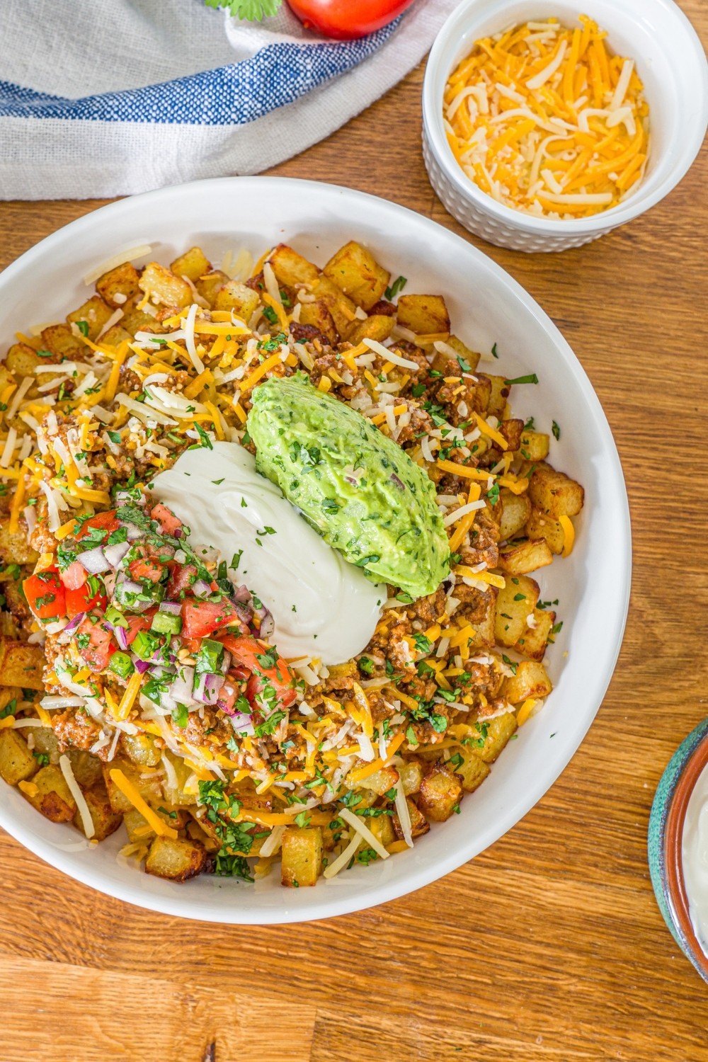 A white bowl with a loaded potato taco bowl including roasted potatoes, taco meat, shredded cheese, pico de gallo, sour cream, and guacamole. The bowl is garnished with cilantro. The bowl is on a wooden counter with a white striped napkin.