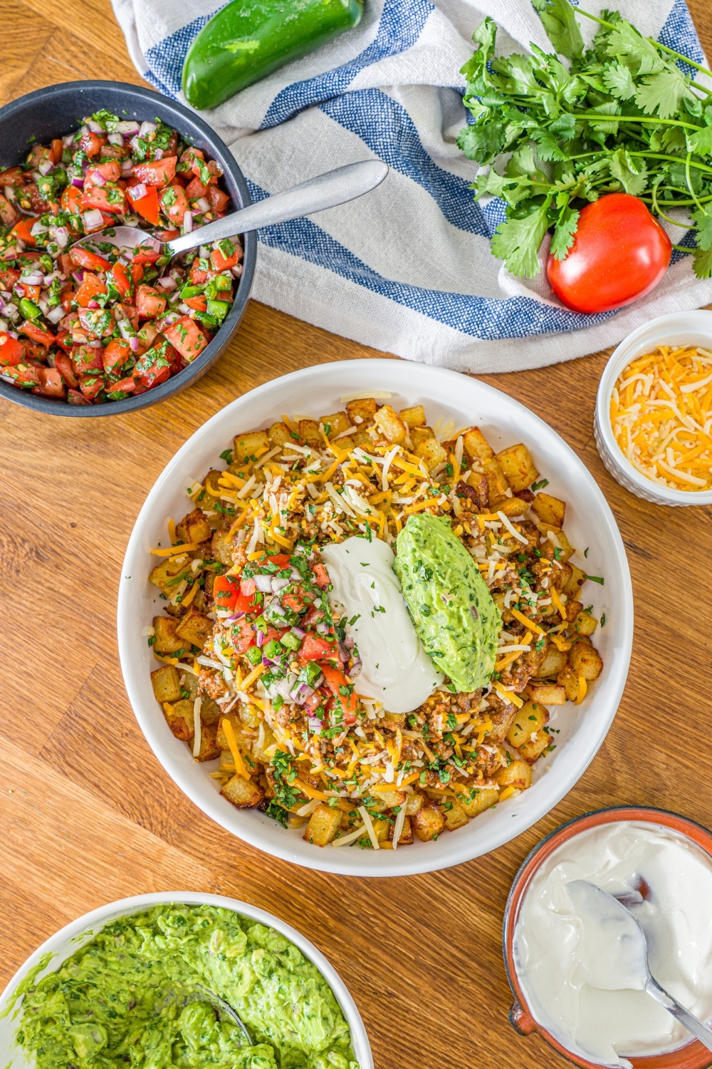 A white bowl with a loaded potato taco bowl including roasted potatoes, taco meat, shredded cheese, pico de gallo, sour cream, and guacamole. The bowl is garnished with cilantro. The bowl is on a wooden counter with bowls of toppings and a white striped napkin.