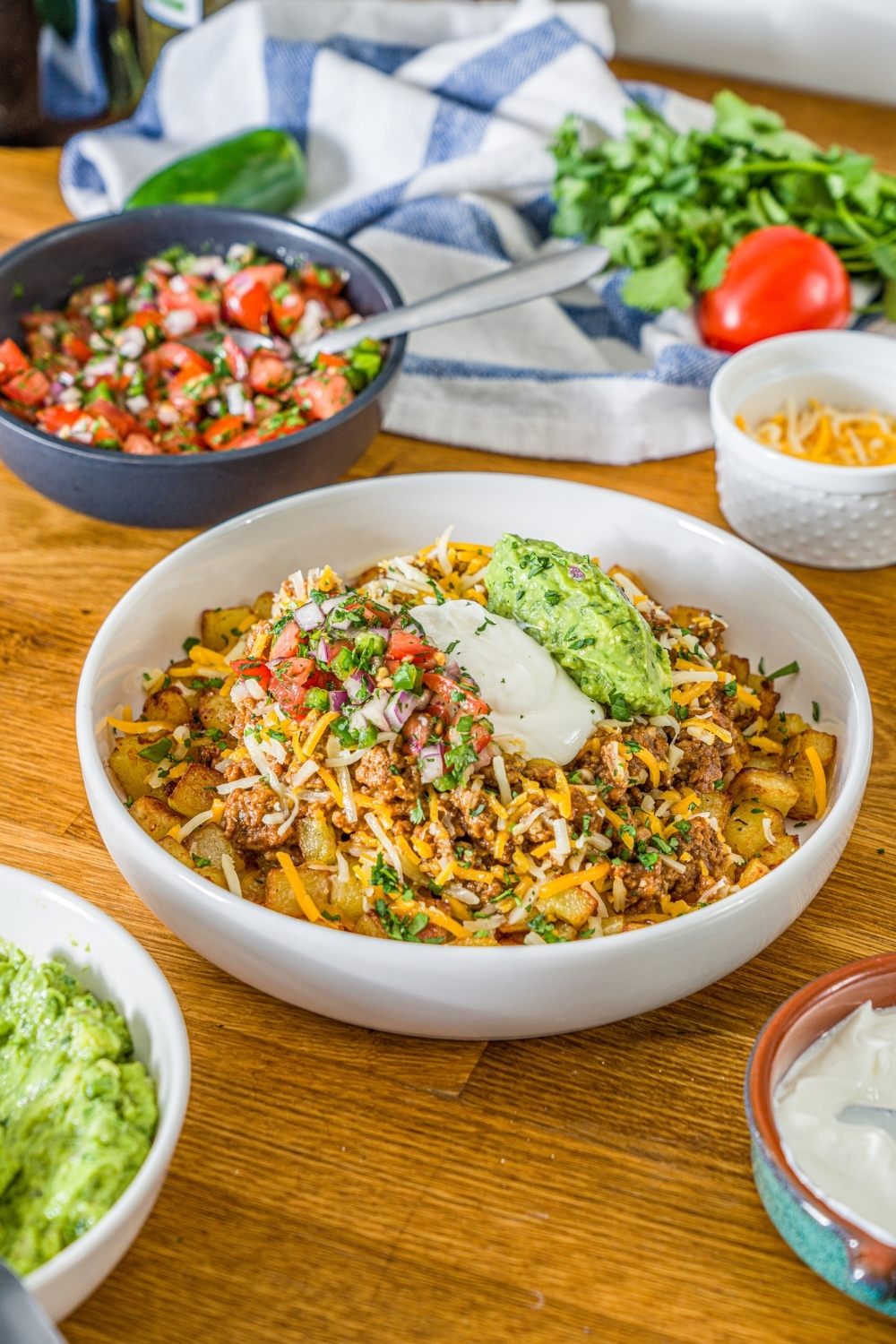 A white bowl with a loaded potato taco bowl including roasted potatoes, taco meat, shredded cheese, pico de gallo, sour cream, and guacamole. The bowl is garnished with cilantro. The bowl is on a wooden counter with bowls of toppings and a white striped napkin.