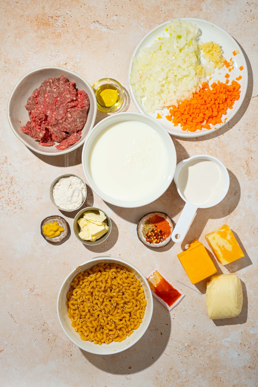 A bowl of ground beef, a plate of onions and carrots, a bowl of cream, three different cheese blocks, a bowl of pasta noodles, a bowl, paprika, and a bowl of butter, all on a white counter.