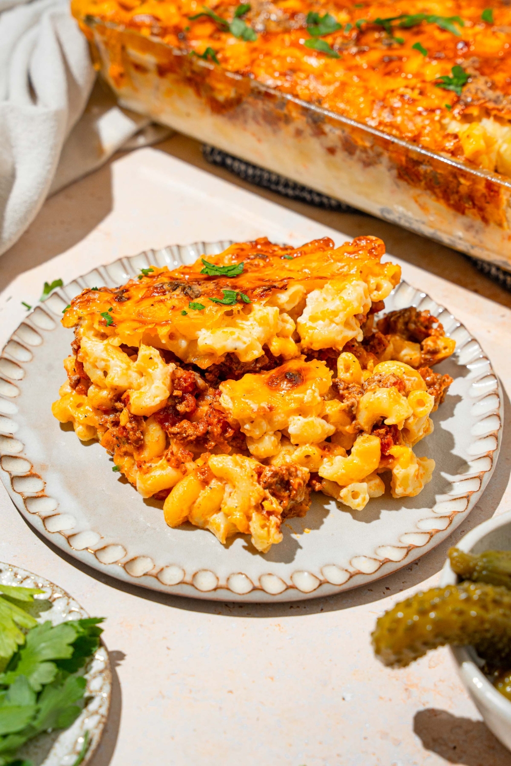 Mac & cheese lasagna on a white plate. Behind it is part of the glass baking dish that has the mac & cheese lasagna in it.