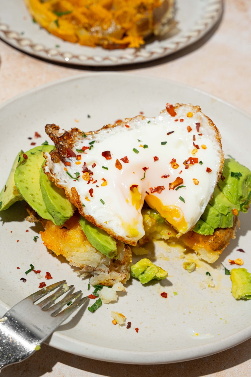 A sunny side up egg on top of smashed avocado on a potato waffle that's all on a white plate.