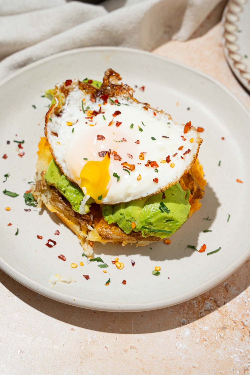 A runny sunny side up egg on top of avocado on a potato waffle on a white plate.