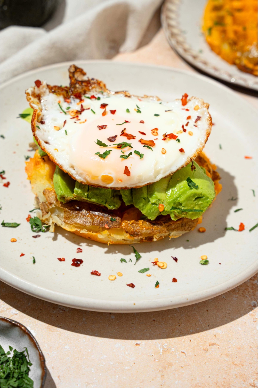 A fried egg on top of sliced avocado on a potato waffle on a white plate.