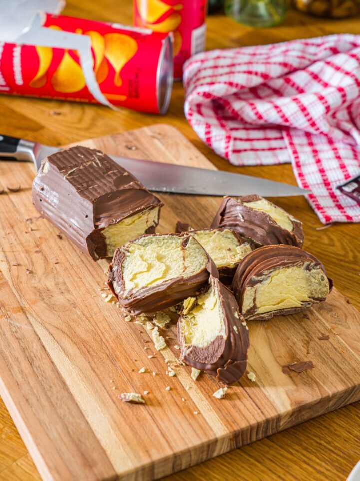 A few pieces of chocolate covered Pringles in front of the Pringles chocolate black all on a wooden cutting board.